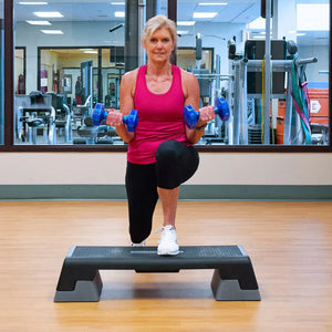 Woman exercising with blue dumbbells on a step platform in a gym.